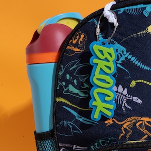 May include: A blue and green name tag that says "BROCK" hangs from a backpack. The backpack is navy blue with a dinosaur skeleton pattern. A blue water bottle with a red and yellow lid is in the side pocket.