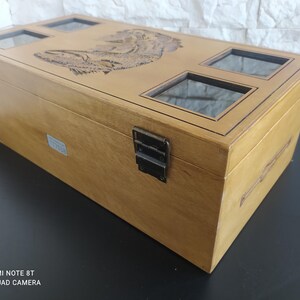 May include: A light brown wooden box with a carved lid design and four glass viewing windows. The box has metal clasps and hinges. The rectangular box is a storage container.