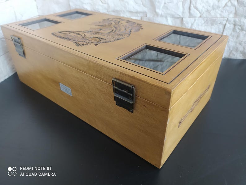 May include: A light brown wooden box with a carved lid design and four glass viewing windows. The box has metal clasps and hinges. The rectangular box is a storage container.