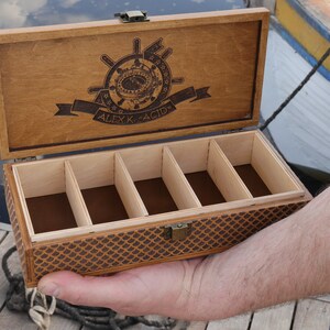 Op de afbeelding: Een houten doos met een scharnierend deksel en een bruine viltvoering. De doos heeft vijf compartimenten en is versierd met een nautisch ontwerp en de tekst "ALEXK. ACIO".