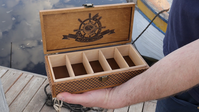 Op de afbeelding: Een houten doos met een scharnierend deksel en een bruine viltvoering. De doos heeft vijf compartimenten en is versierd met een nautisch ontwerp en de tekst "ALEXK. ACIO".