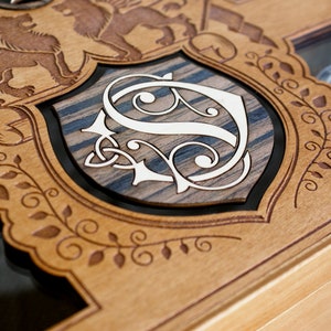 May include: A wooden box with a decorative design featuring a shield with a stylized letter "S" and two winged lions. The box has a glass top and is designed to hold watches.