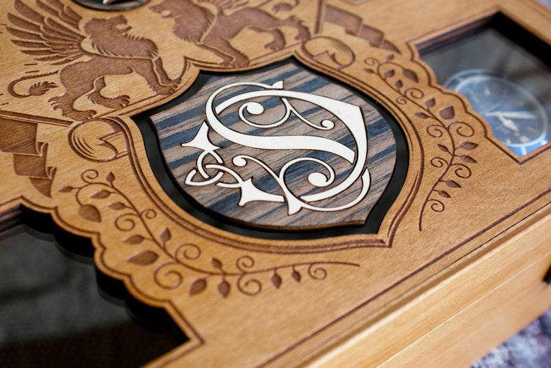 May include: A wooden box with a decorative design featuring a shield with a stylized letter "S" and two winged lions. The box has a glass top and is designed to hold watches.