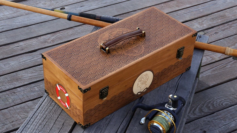 May include: A rectangular wooden tackle box with a brown leather handle, brass-colored metal corner accents, and two front latches. The lid features a laser-engraved wave pattern, a circular life preserver graphic, and a side portrait silhouette.