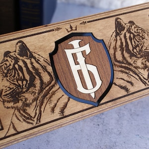 May include: A wooden watch box with two tiger engravings, a shield with the initials "F.S." and a crown, and two watch compartments. The box is made of light brown wood with a dark brown stain.