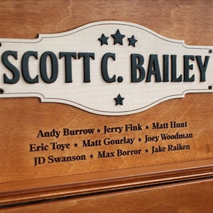 K&ouml;nnte beinhalten: Eine Holzplakette mit wei&szlig;em Hintergrund und schwarzer Beschriftung. Die Plakette tr&auml;gt die Aufschrift "SCOTT C. BAILEY" mit drei Sternen &uuml;ber dem Namen und einem Stern darunter. Unter der Plakette sind die Namen mehrerer Personen in schwarzer Schrift aufgef&uuml;hrt.