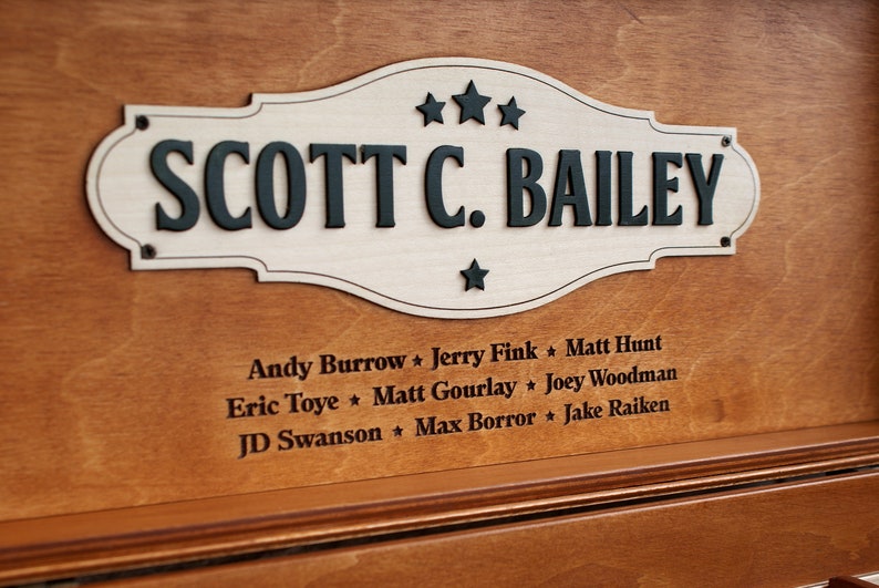 K&ouml;nnte beinhalten: Eine Holzplakette mit wei&szlig;em Hintergrund und schwarzer Beschriftung. Die Plakette tr&auml;gt die Aufschrift "SCOTT C. BAILEY" mit drei Sternen &uuml;ber dem Namen und einem Stern darunter. Unter der Plakette sind die Namen mehrerer Personen in schwarzer Schrift aufgef&uuml;hrt.
