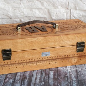 May include: Wooden box with a handle and metal hardware. The box has a ruler etched into the front and decorative engravings on the top. The initials "AB" are displayed on the top. The box has a brown leather handle.