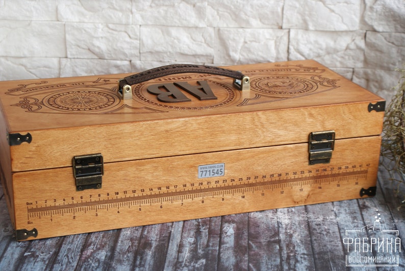 May include: Wooden box with a handle and metal hardware. The box has a ruler etched into the front and decorative engravings on the top. The initials "AB" are displayed on the top. The box has a brown leather handle.