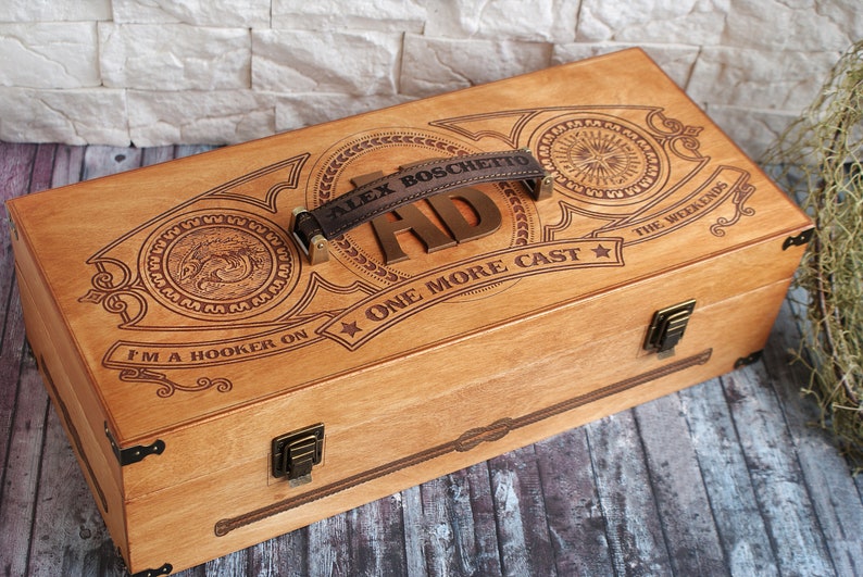 May include: Wooden fishing tackle box with engraved details. Features a leather handle and metal clasps. The box is decorated with the text "ALEX BOSCHETTO HD ONE MORE CAST" and "I'M A HOOKER ON THE WEEKENDS".