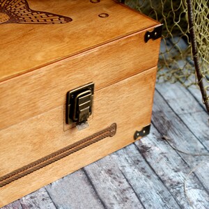 May include: A wooden box with a fish design and a brass latch. The box is on a wooden surface with a fishing net in the background.