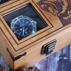 May include: A wooden watch box with a glass top, featuring a blue-faced watch inside. The box has a brass latch and is decorated with an engraved design of a bear's head.