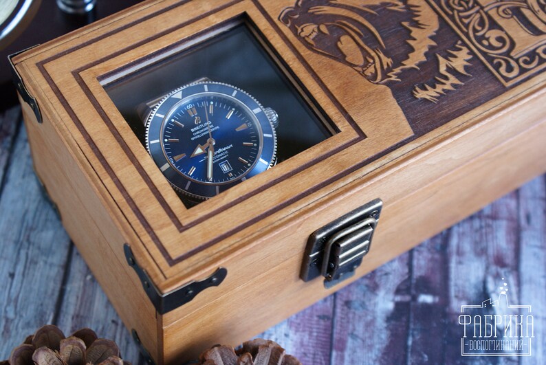 May include: A wooden watch box with a glass top, featuring a blue-faced watch inside. The box has a brass latch and is decorated with an engraved design of a bear's head.