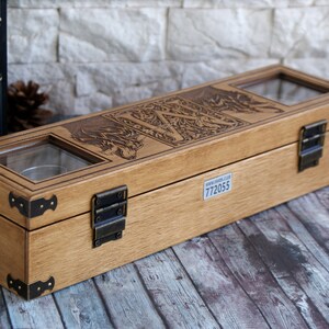 May include: A wooden box with a hinged lid and brass accents. The box has a glass window on the top and is engraved with a design of a wolf and the letter "W". The box has a label on the bottom with the number "772055".