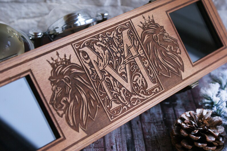 Pu&ograve; includere: Una scatola di legno con un coperchio incernierato, che presenta un design inciso di una lettera stilizzata "M" circondata da motivi floreali decorativi. Due leoni con corone sono incisi su entrambi i lati della lettera. La scatola ha due piccole finestre rettangolari sul davanti.