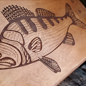 May include: A wooden box with a laser-engraved image of a fish. The box is light brown and the fish is dark brown. The text "Фабрика" and "водоочистки" is printed on the bottom of the box.