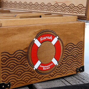 May include: A wooden storage chest with a hinged lid, featuring a laser-engraved wave pattern and a central red and white life ring graphic. The life ring displays the name Sarah and the text Bridgehampton, New York. It has metal corner accents.