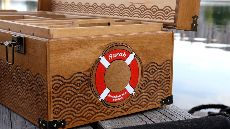 May include: A wooden storage chest with a hinged lid, featuring a laser-engraved wave pattern and a central red and white life ring graphic. The life ring displays the name Sarah and the text Bridgehampton, New York. It has metal corner accents.