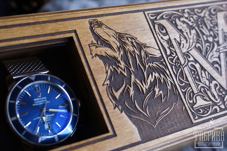 May include: A wooden watch box with a blue watch inside. The box is engraved with a wolf and the letter "M" in a decorative font. The text "Фабрика - воплощения" is also engraved on the box.