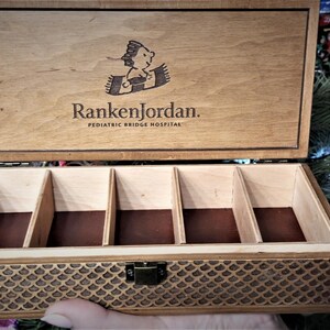 Op de afbeelding: Een houten doos met een scharnierend deksel en een compartimenterend interieur. Het deksel is gegraveerd met de tekst "Ranken Jordan. Pediatric Bridge Hospital" en een cartoonafbeelding van een kind op een tapijt. De doos heeft een decoratief patroon op de bodem.