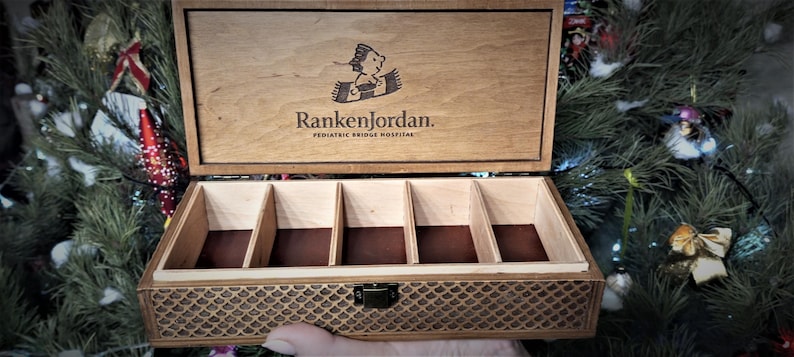 Op de afbeelding: Een houten doos met een scharnierend deksel en een compartimenterend interieur. Het deksel is gegraveerd met de tekst "Ranken Jordan. Pediatric Bridge Hospital" en een cartoonafbeelding van een kind op een tapijt. De doos heeft een decoratief patroon op de bodem.