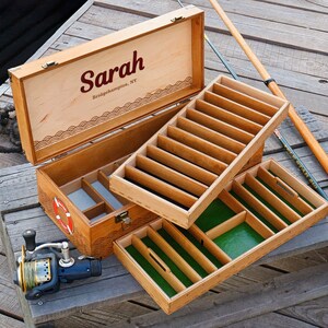 May include: A wooden fishing tackle box with a hinged lid featuring the name Sarah and Bridgehampton, NY. The interior has removable trays with green felt lining and dividers. A fishing reel and rod are positioned next to the open box.