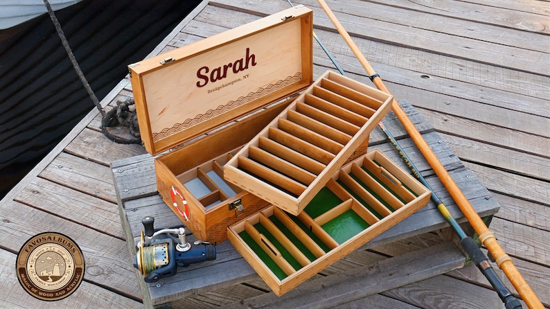 May include: A wooden fishing tackle box with a hinged lid featuring the name Sarah and Bridgehampton, NY. The interior has removable trays with green felt lining and dividers. A fishing reel and rod are positioned next to the open box.