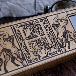 May include: A wooden watch box with a decorative design featuring a stylized letter "H" surrounded by floral motifs and two bulls. The box has a glass window displaying a watch face.