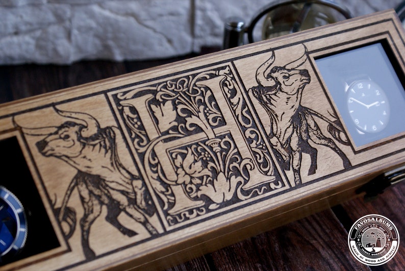 May include: A wooden watch box with a decorative design featuring a stylized letter "H" surrounded by floral motifs and two bulls. The box has a glass window displaying a watch face.