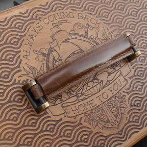 May include: A rectangular wooden box lid featuring a laser-engraved wave pattern and a central illustration of a ship with the text ALWAYS COMING BACK and HOME TO YOU. A brown leather handle with brass-colored metal hardware is attached to the center.
