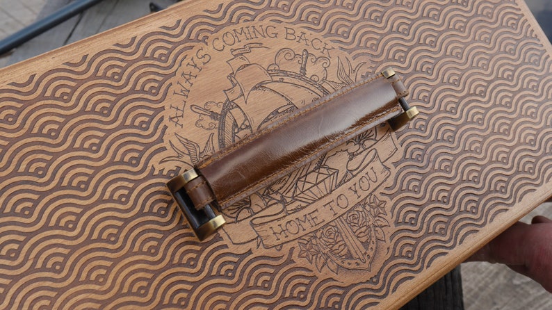 May include: A rectangular wooden box lid featuring a laser-engraved wave pattern and a central illustration of a ship with the text ALWAYS COMING BACK and HOME TO YOU. A brown leather handle with brass-colored metal hardware is attached to the center.