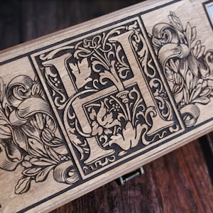 May include: A wooden box with a hinged lid, featuring an intricate laser-engraved design of a stylized letter "H" surrounded by floral motifs. The box has a dark brown stain and a clear glass window on the right side.