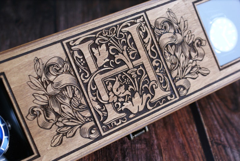 May include: A wooden box with a hinged lid, featuring an intricate laser-engraved design of a stylized letter "H" surrounded by floral motifs. The box has a dark brown stain and a clear glass window on the right side.