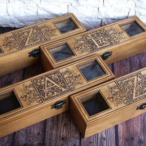 May include: Five wooden boxes with glass lids, each featuring a different letter of the alphabet in a decorative, ornate design. The boxes are arranged in a staggered pattern on a wooden surface.