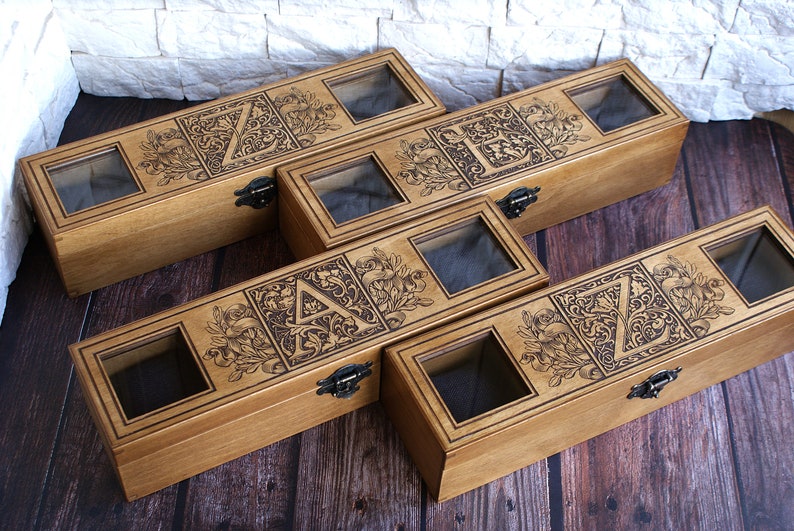 May include: Five wooden boxes with glass lids, each featuring a different letter of the alphabet in a decorative, ornate design. The boxes are arranged in a staggered pattern on a wooden surface.