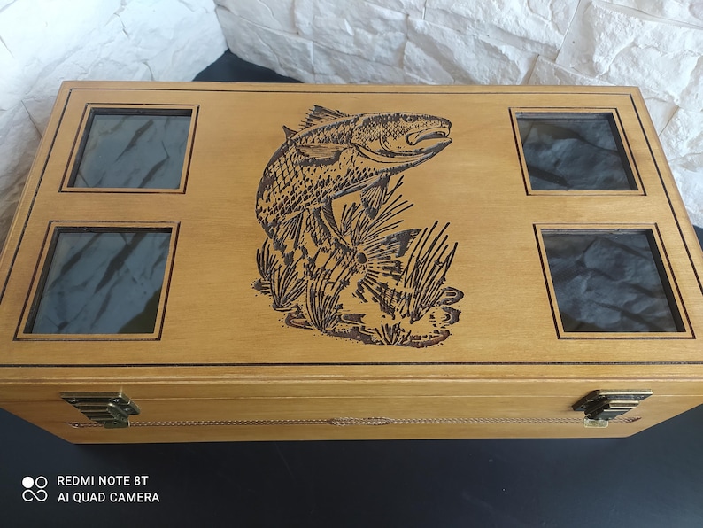 May include: A light-coloured wooden box with four glass-covered compartments and a hinged lid. The box features a detailed engraving of a fish leaping from the water, surrounded by reeds. It has metal clasps.