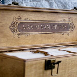 May include: A wooden box with a hinged lid and a decorative engraved inscription that reads "MAXIM VAN OPPENS". The box has a latch and is divided into compartments.