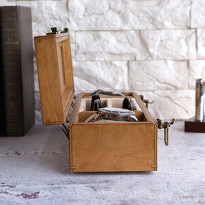 Peut inclure: Une bo&icirc;te de rangement pour montres en bois rectangulaire avec un couvercle &agrave; charni&egrave;re et un loquet en m&eacute;tal, dot&eacute;e de compartiments internes doubl&eacute;s de tissu beige. La bo&icirc;te est ouverte, r&eacute;v&eacute;lant une montre-bracelet argent&eacute;e &agrave; l'int&eacute;rieur.