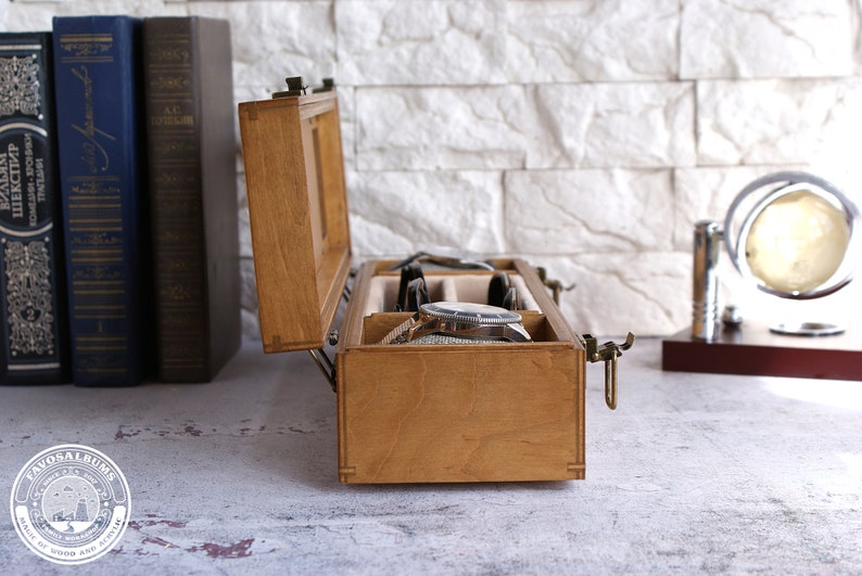 Peut inclure: Une bo&icirc;te de rangement pour montres en bois rectangulaire avec un couvercle &agrave; charni&egrave;re et un loquet en m&eacute;tal, dot&eacute;e de compartiments internes doubl&eacute;s de tissu beige. La bo&icirc;te est ouverte, r&eacute;v&eacute;lant une montre-bracelet argent&eacute;e &agrave; l'int&eacute;rieur.