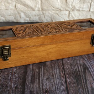 May include: A wooden box with a hinged lid and two brass clasps. The box has a carved design on the top and three glass windows on the lid. The box is sitting on a dark wooden surface.