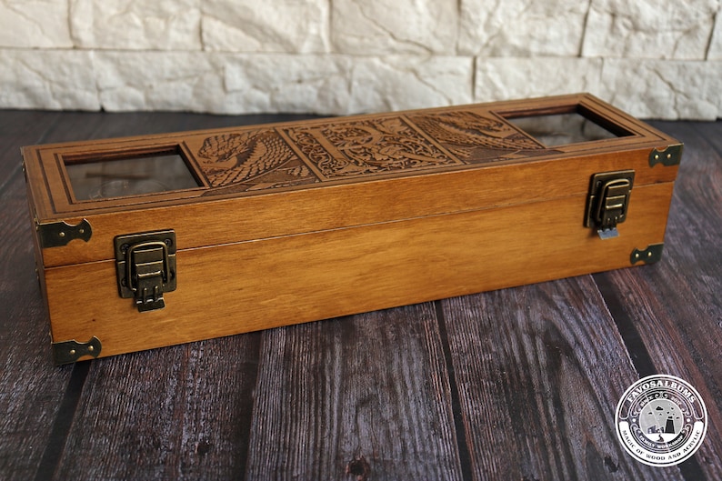 May include: A wooden box with a hinged lid and two brass clasps. The box has a carved design on the top and three glass windows on the lid. The box is sitting on a dark wooden surface.