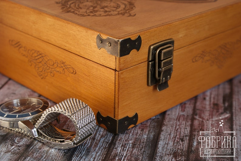 K&ouml;nnte beinhalten: Eine Holzkiste mit einem Messingverschluss und dekorativen Metall-Ecken. Die Kiste ist geschlossen und ein Uhrenarmband liegt auf einer Holzoberfl&auml;che davor.