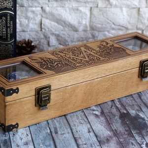 May include: A wooden box with two glass windows, featuring an engraved design of a wolf and the letter "M". The box has two brass clasps and is sitting on a wooden surface.