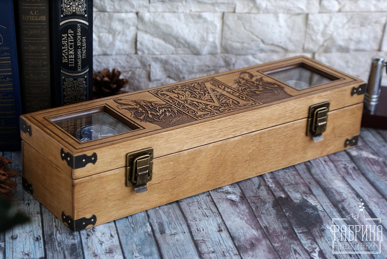 May include: A wooden box with two glass windows, featuring an engraved design of a wolf and the letter "M". The box has two brass clasps and is sitting on a wooden surface.