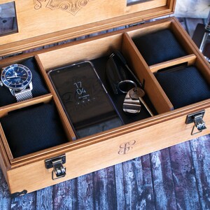 Puede incluir: Una caja de madera con tapa abatible, que contiene compartimentos para guardar relojes, un tel&eacute;fono y otros objetos peque&ntilde;os. La caja tiene un cierre y est&aacute; hecha de madera marr&oacute;n claro. La caja tiene una inscripci&oacute;n personalizada en la parte delantera.