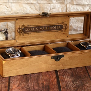 May include: A wooden watch box with a clear glass top and four compartments. The box is engraved with the name "MARTIN MAIERHOFER".