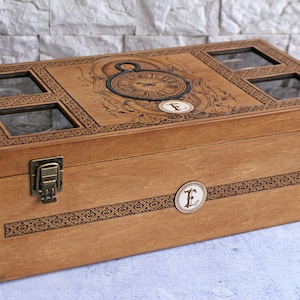 May include: A wooden watch box with a decorative clock face design and four compartments with glass lids. The box has brass clasps and a personalized engraved letter "E" on the front.