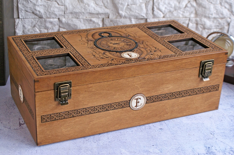 May include: A wooden watch box with a decorative clock face design and four compartments with glass lids. The box has brass clasps and a personalized engraved letter "E" on the front.