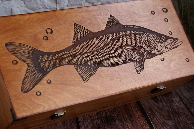 K&ouml;nnte beinhalten: Eine Holzkiste mit einem lasergravierten Bild eines Fisches. Der Fisch ist detailliert mit Schuppen und Flossen dargestellt. Die Kiste ist braun gebeizt und hat zwei Messing Scharniere.