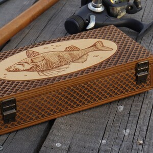 Op de afbeelding: Een houten doos met een visontwerp op de deksel. De doos heeft een bruine afwerking en een schubbenpatroon aan de zijkanten. De doos heeft twee messing sluitingen.